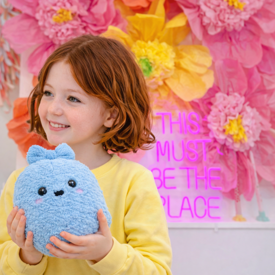 Child holding a blue plushie toy hand knitted in front of colourful floral paper decorations and a neon sign saying "This must be the place"