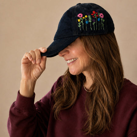 A woman wearing a dark navy blue denim baseball cap with colourful embroidered flowers on the front, paired with a burgundy sweatshirt against a solid beige background.