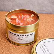 Handmade soy candle in a gold tin with floral elements on a textured surface.