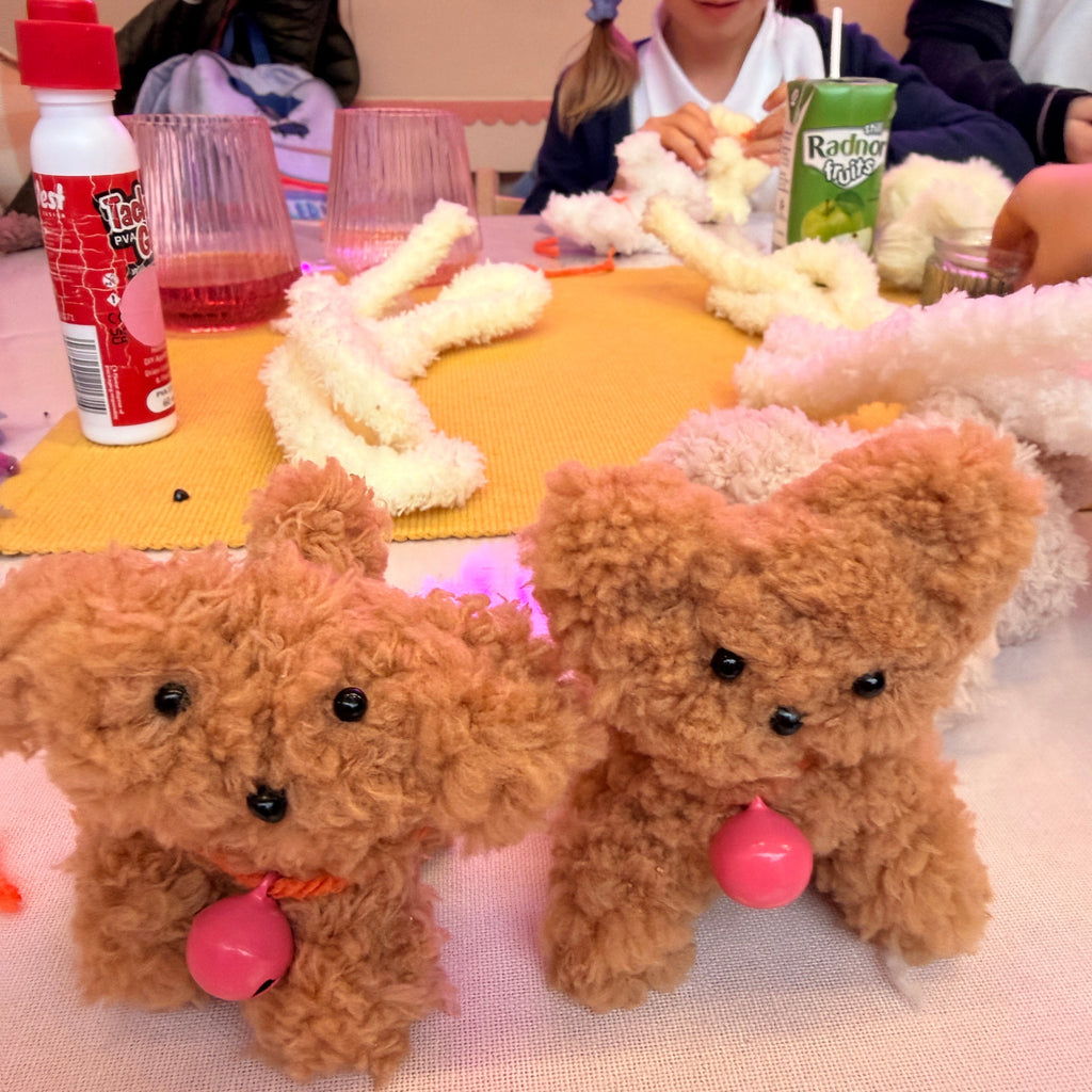 Two small plush toy puppy dogs with curly brown fur and pink bellies are arranged on a table, alongside craft supplies including glue, scissors, and a green juice box. A person is partially visible in the background.