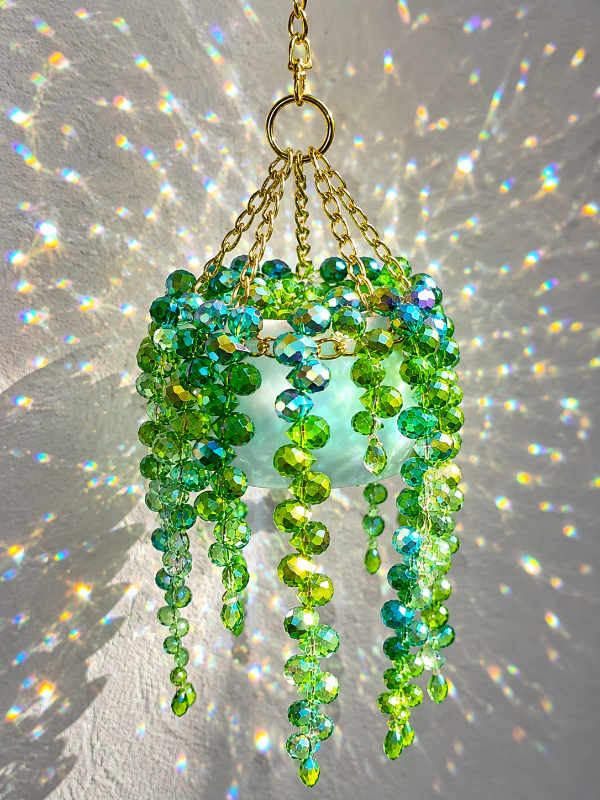A hanging chandelier with a gold-tone chain and ring, decorated with cascading strands of faceted green beads and iridescent blue-green beads, shown against a white wall with colorful light reflections.