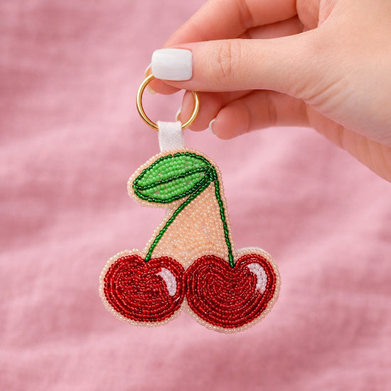 Hand holding a beaded cherry keychain against a pink background