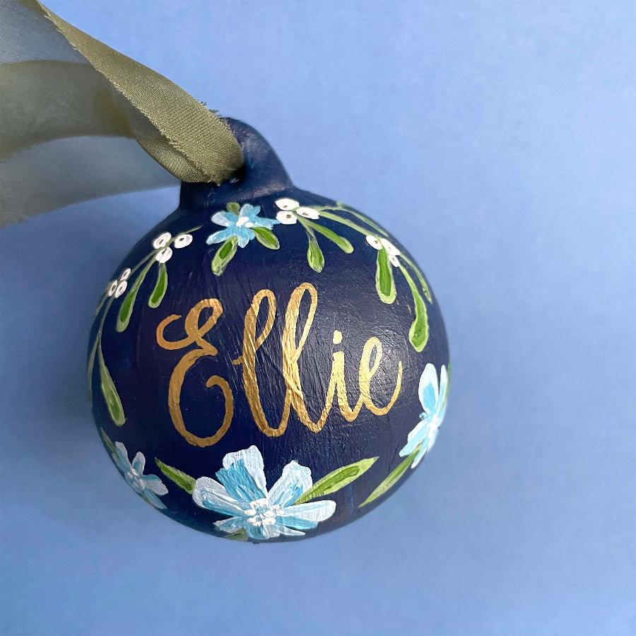 A personalized dark blue Christmas ornament with hand-painted light blue and white flowers, green leaves, and gold foil script reading 'Ellie', hanging from a gold ribbon against a light blue background.